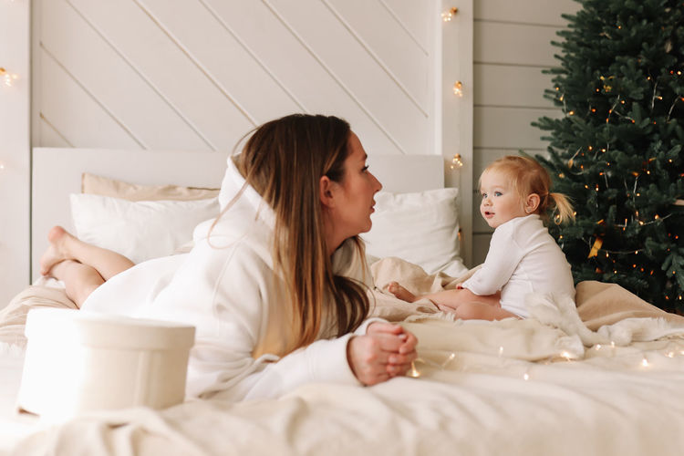 Moody sad little daughter child offended by mom sitting on the bed at the christmas tree at home