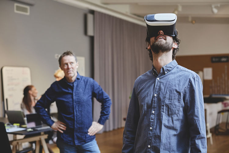 Mature businessman standing with hands on hip while looking at male colleague using virtual reality simulator in creativ