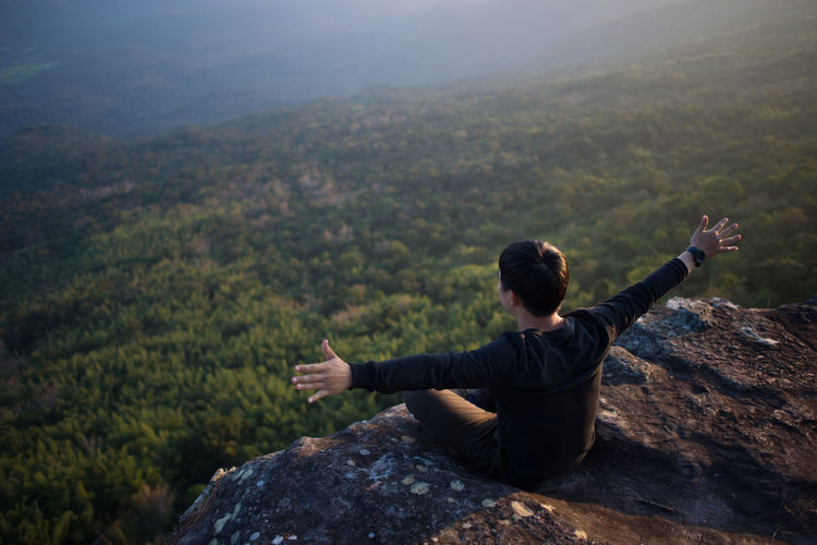 High angle view of young man with arms outstretched sitting on rock