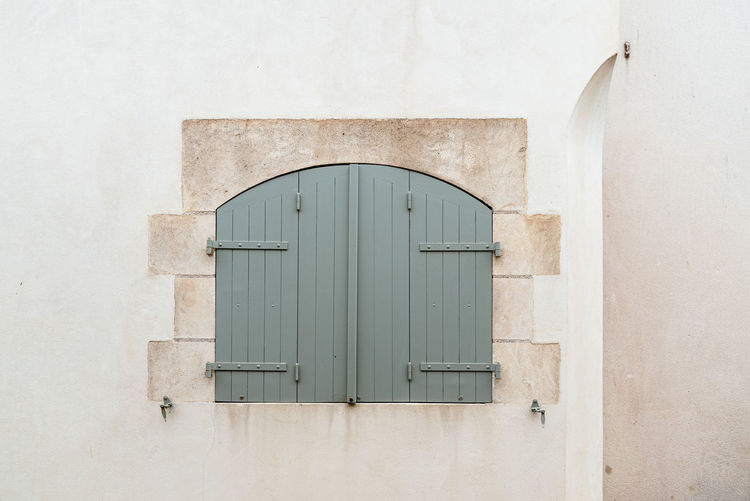 Old window with wooden light green painted closed shutters on white wall