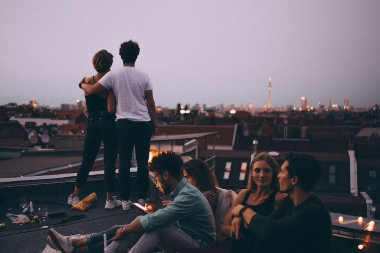 Couple looking at city while friends relaxing on terrace during rooftop party