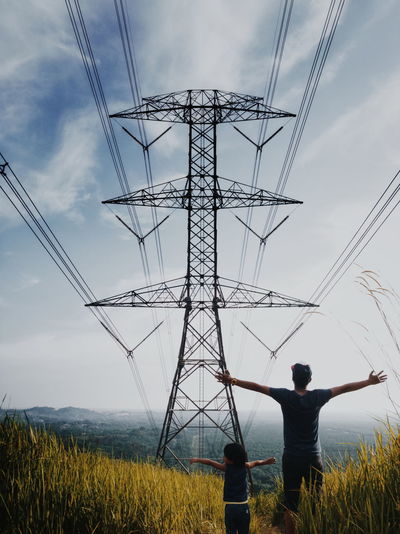 Rear view of man by daughter standing with arms outstretched by pylon on field