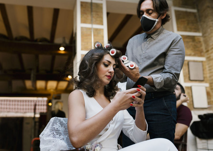 Male hairdresser doing hairstyle while bride using smart phone at salon during pandemic