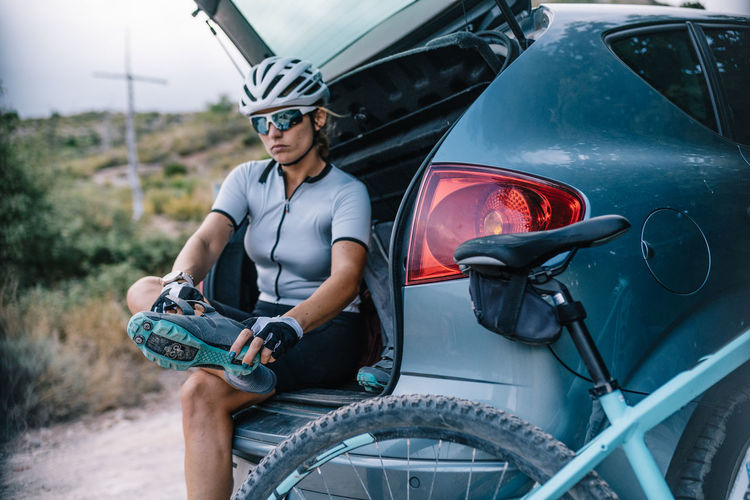 Confident female bicyclist in helmet and sunglasses sitting in trunk of car and preparing for ride while putting on boots and looking at camera