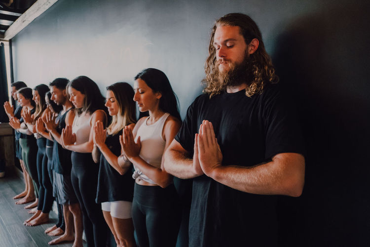 Group of happy young sporty people in activewear with eyes closed relaxing and deep breathing while standing with namaste in tadasana and meditating beside black wall in modern yoga studio