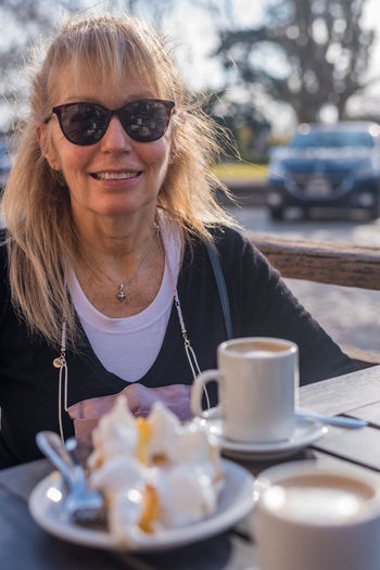 Mature adult woman smiling wearing glasses with a latte and lemon pie. looking at the camera