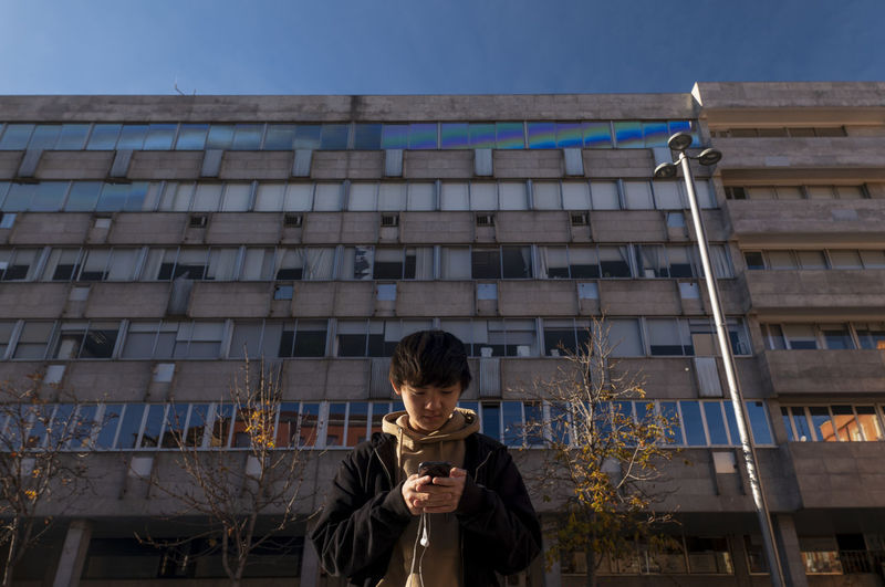Asian teenage boy looking at mobile phone against building on street. madrid. spain