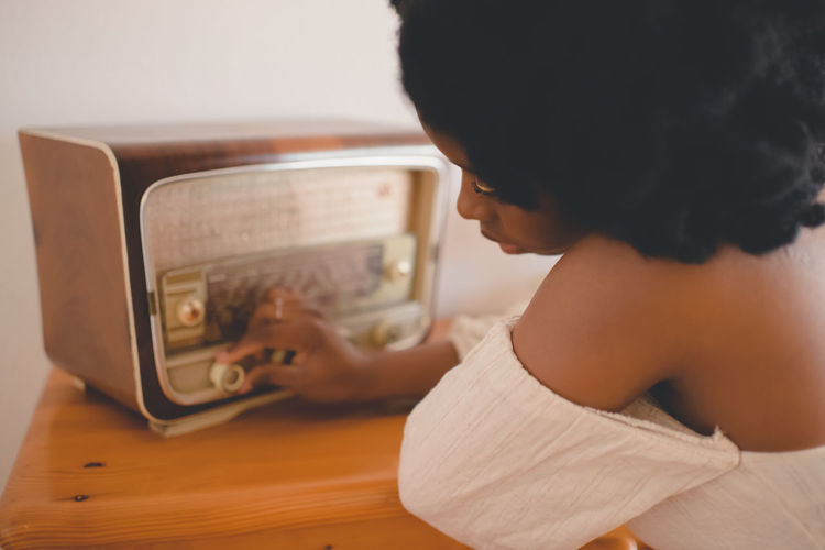 Side view of young african american female sitting at table and tuning old fashioned radio receiver while resting at home