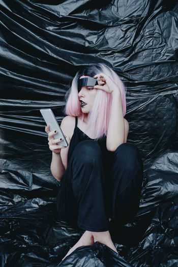 Woman with futuristic eyeglasses using smart phone sitting on black plastic backdrop