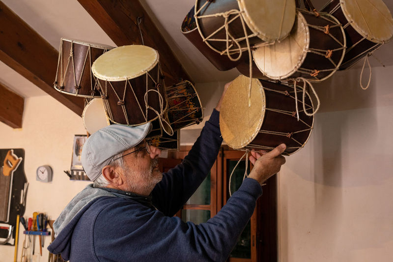 Side view of focused male master in cap hanging traditional tapan drums with lacing on ceiling in professional light workshop