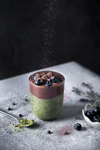 Glass of appetizing healthy matcha tea and chia seeds smoothie with chocolate and blueberries sprinkled with cocoa powder on dusted with sugar powder table