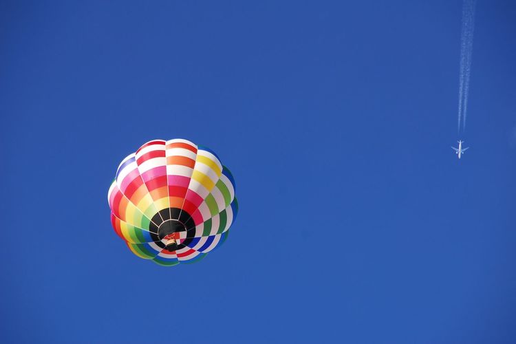Low angle view of vivid colored balloon and airliner with contrail in winter blue sky