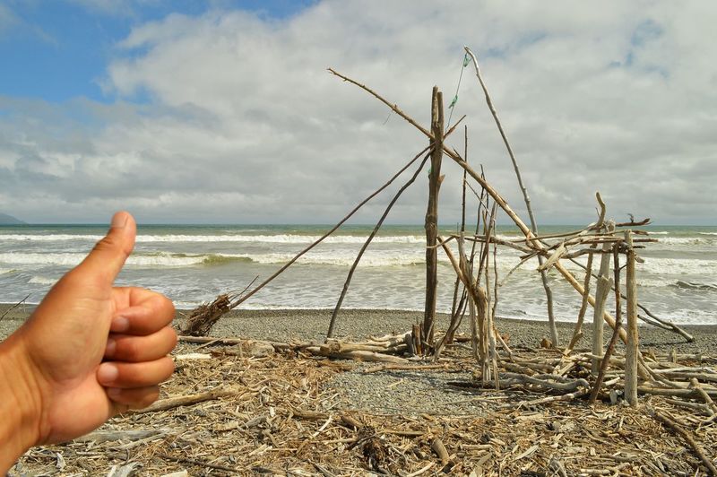 Human hand forming ok sign before broken wooden structure on beach against cloudy sky