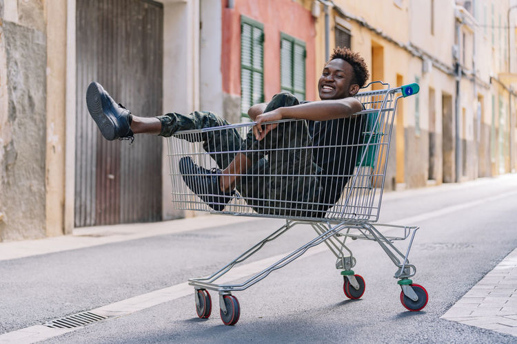 Side view of pensive youthful african american male teenager in casual clothes looking at camera while sitting in metal shopping trolley in street