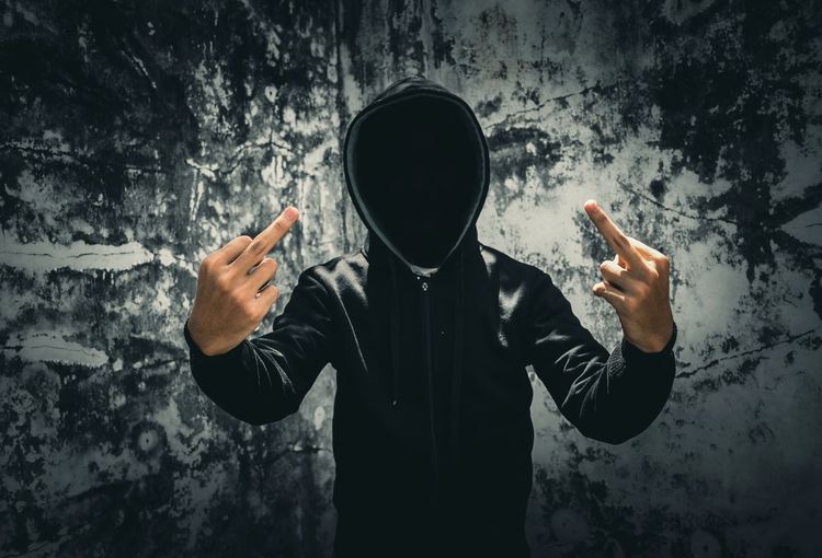 Man in hooded jacket showing middle finger while standing against old wall