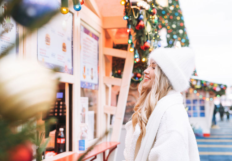 Young happy woman with curly hair in white knitted hat on shopping at christmas fair market 