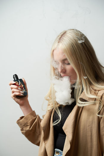 Tranquil female in stylish outfit standing in studio and smoking vape while looking down