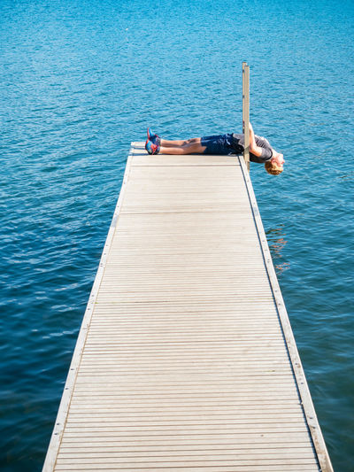 Blond boy lying on wooden pier over lake and staring at the blue sky above him. boy enjoys childhood