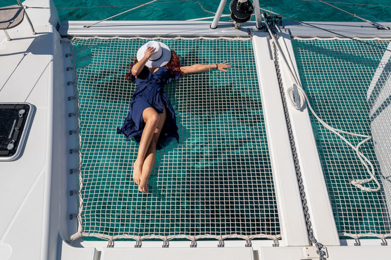 From above of unrecognizable female in dress and sunhats lying down on net of sailing yacht on sunny day during summer holiday