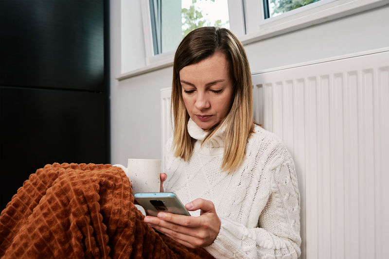 Worried woman sit near heating radiator under blanket and using smartphone