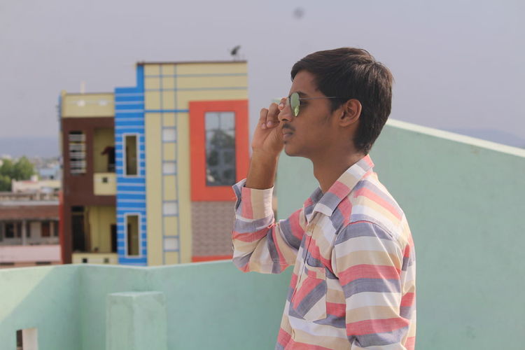 Side view of young man holding sunglasses while standing on building terrace