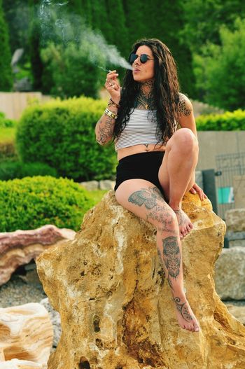 Full length of woman smoking cigarette while sitting on rock
