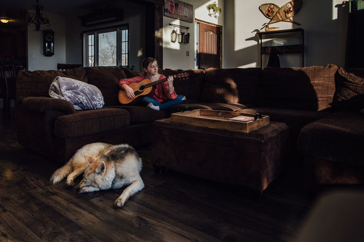 Young girl playing guitar on couch with dog sleeping nearby