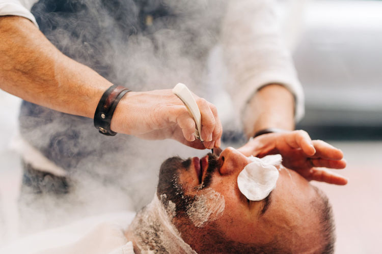 Crop anonymous beauty master shaving beard of client with straight razor during steam vapor treatment in hairdressing salon