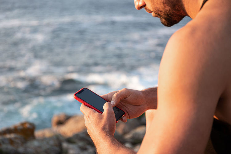 Side view of male in summer clothes sitting on rock near sea and messaging on social media via cellphone at sunset