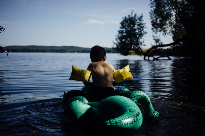 Rear view of boy sitting on inflatable crocodile at lake against sky
