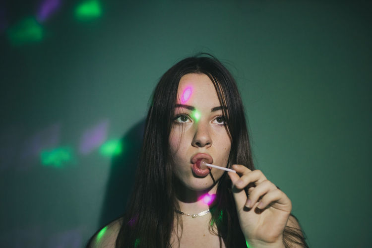 Young woman licking lollipop while lights falling on her against green background