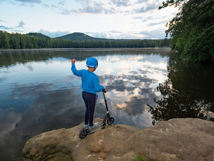 Teenager boy in a blue helmet is stand at lake within sunset against a blue sky. with a push scooter