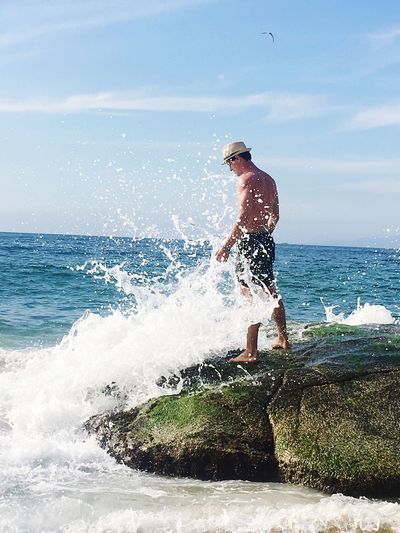 Sea waves splashing on shirtless man standing on rock against sky