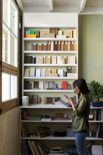 Craftswoman looking at wooden samples while standing by shelf in workshop