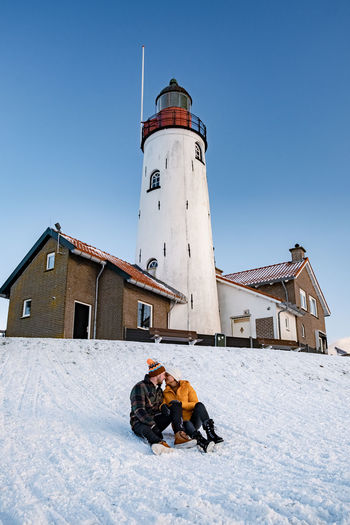 Full length of couple sitting on snow covered land against lighthouse