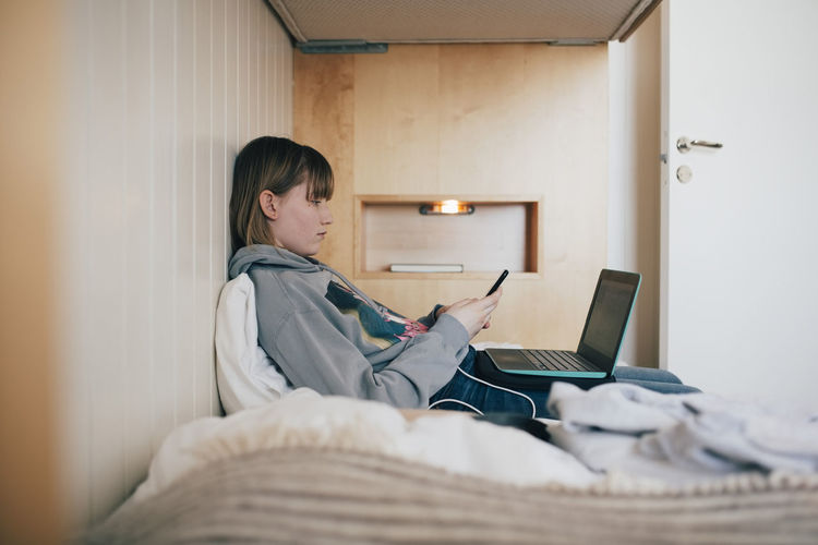 Teenage girl sitting with laptop using smart phone in vacation home