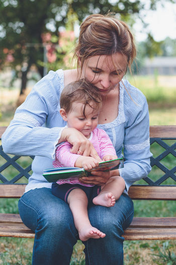 Mother showing book to daughter while sitting on bench in yard