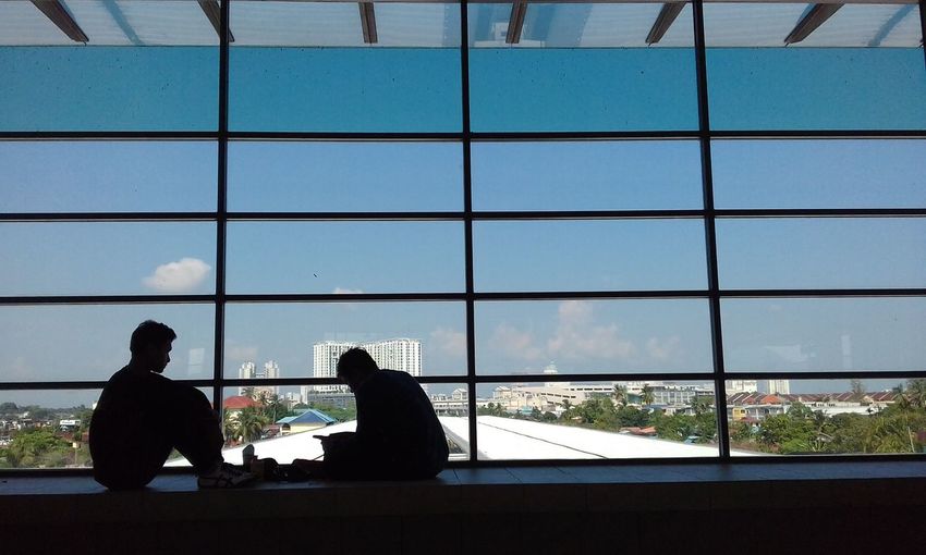 Silhouette men sitting by window in city against sky