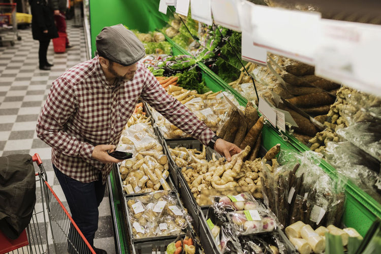 High angle view of man buying root vegetable while holding smart phone in supermarket