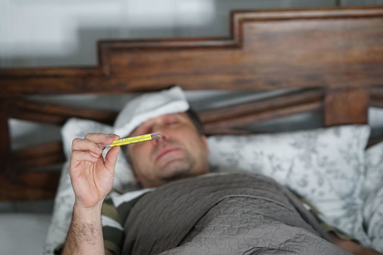 Man holding thermometer while relaxing on bed at home