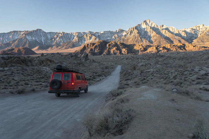 Red camper vehicle driving between fields on long roadway against range of rough rocky cliffs covered with snow on winter day in alabama hills, california, usa