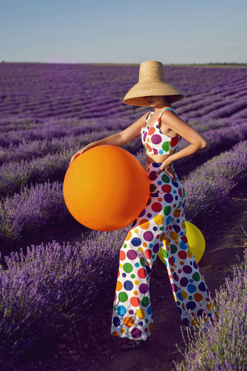 Fashionable young woman in hat on lavender field stands with colored balls and clothes to the point