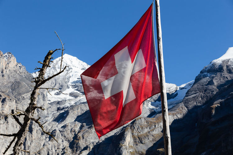 Low angle view of swiss flag by rocky mountains against clear blue sky