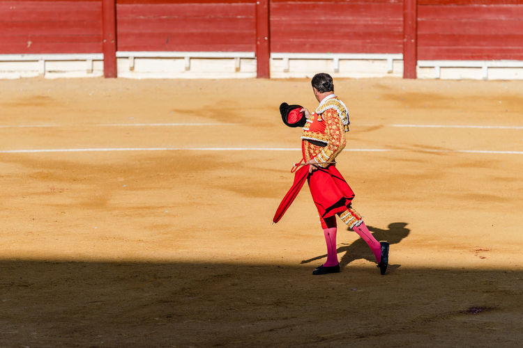 Back view of unrecognizable bullfighter in fancy costume taking off hat after corrida performance while standing on sandy arena
