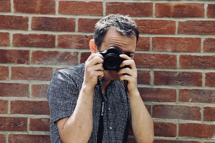 Portrait of man photographing against brick wall