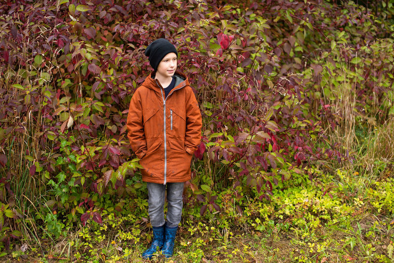 A cute boy in rubber boots and a jacket is standing in the autumn forest