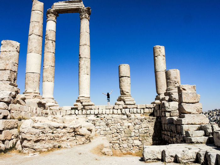 Rear view of woman with arms outstretched standing at amman citadel against clear blue sky