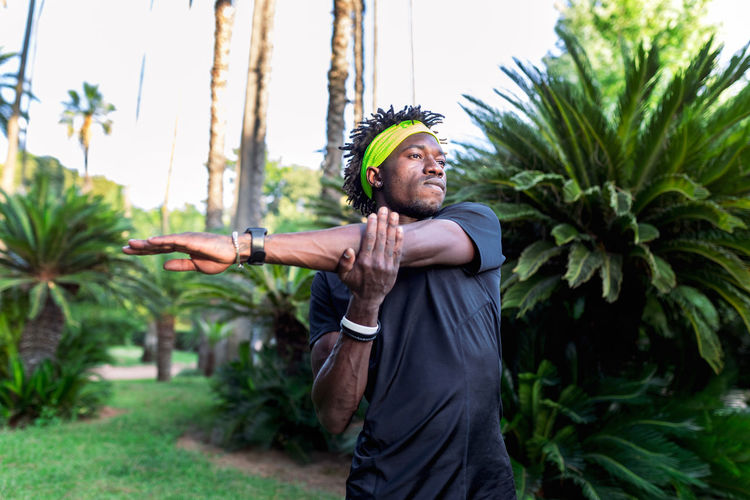 Confident young african american male athlete in trendy sportswear stretching arms while training in park with green tropical trees