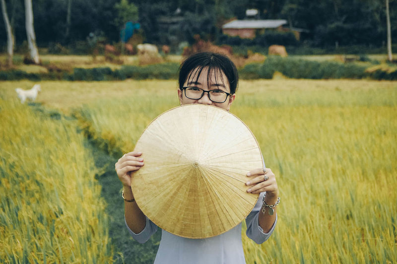 Portrait of woman holding asian style conical hat while standing on field