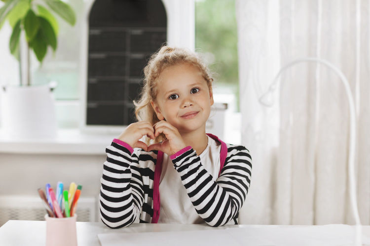 A cute little girl is sitting at a table with a sketchbook and a glass of colored pencils. 
